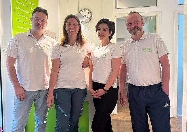 Zwei Therapeutinnen und zwei Therapeuten in weißen Poloshirts stehen nebeneinander in der Praxis.
