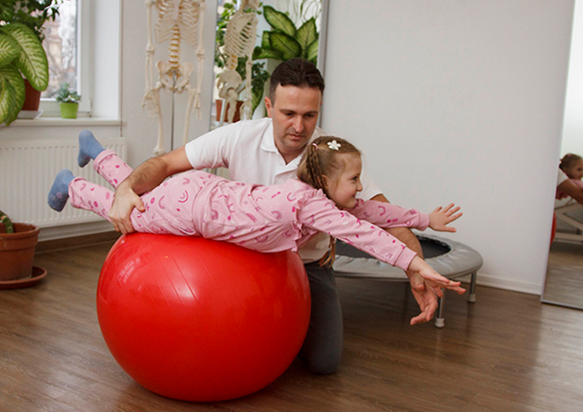 Ein kleines Mädchen liegt in Bauchlage auf einem roten Gymnastikball und streckt die Arme nach vorne aus, während ein Physiotherapeut daneben kniet und das Kind sichert.