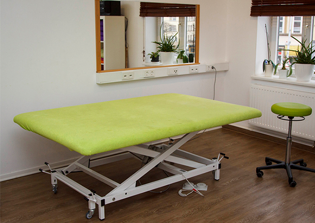 Eine moderne, höhenverstellbare Therapieliege mit hellgrünem Bezug steht in einem hellen Behandlungsraum mit Laminatboden.