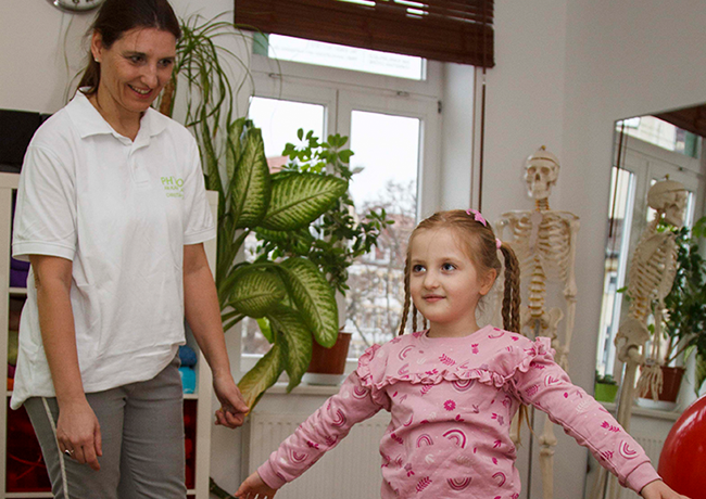 Ein kleines Mädchen im rosa Pullover lächelt bei einer Gleichgewichtsübung, während eine Physiotherapeutin im weißen Poloshirt unterstützend links hinter ihr steht.