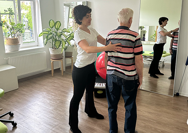 Eine Physiotherapeutin hat ihre Hand sanft auf den Rücken eines älteren Patienten gelegt und korrigiert die aufrechte Körperhaltung.